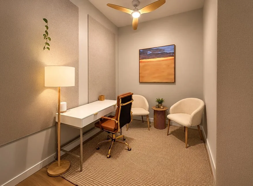 A cozy home office with a white desk, a brown and black swivel chair, two beige armchairs, a small wooden side table with a potted plant, a modern painting of a sunset, a ceiling fan, a floor lamp, and a beige carpet.