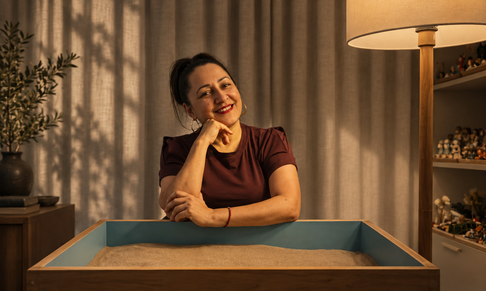 A woman smiling and sitting at a table with a sandbox in front of her, surrounded by an indoor setting with a plant, books, and shelves of figurines.