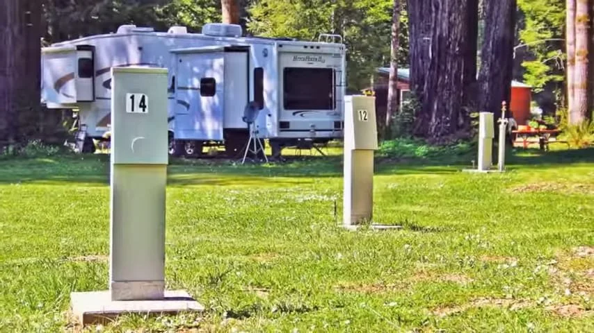 RV parked in a forested campground with electrical hookups and outdoor chairs.