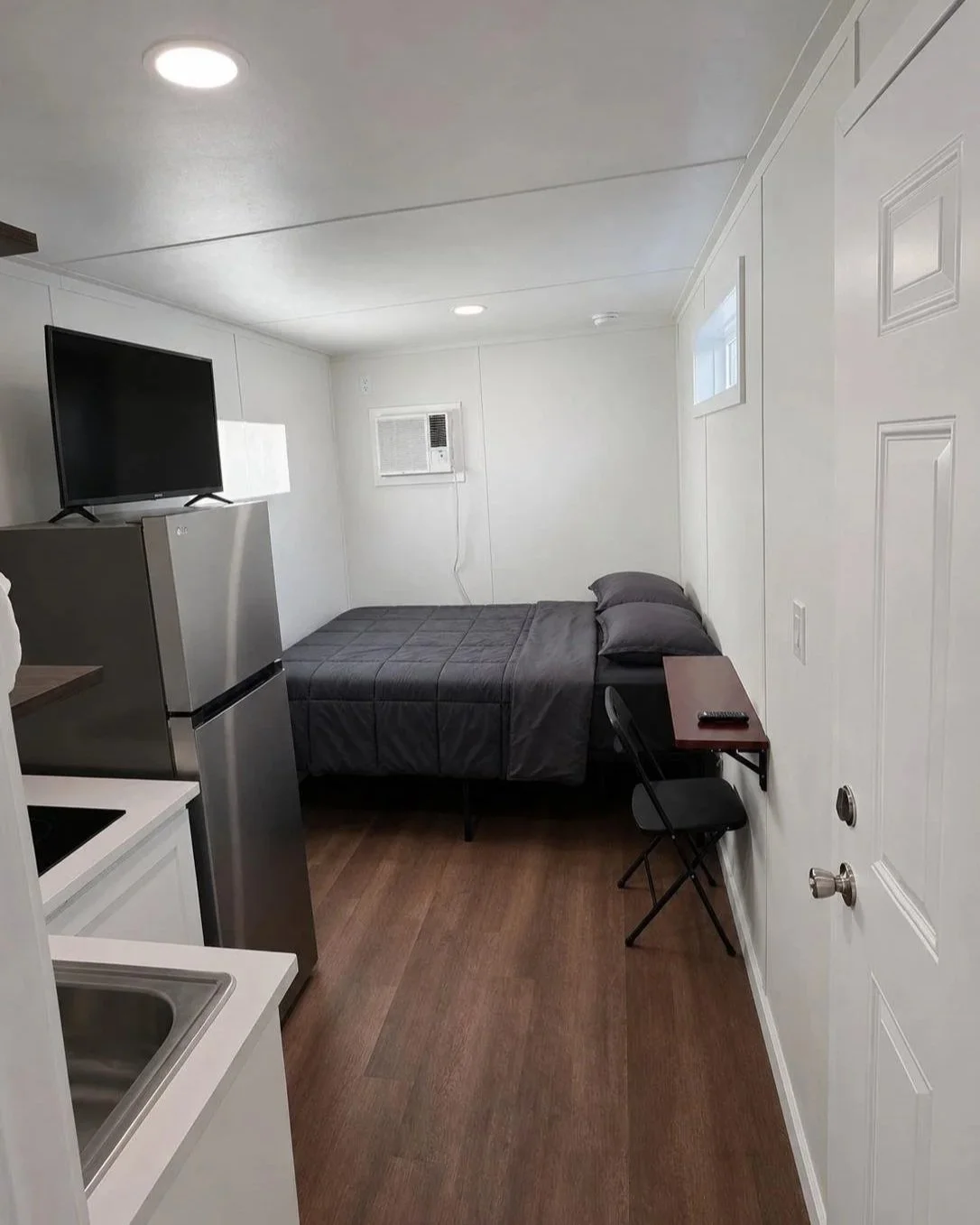 Small bedroom with a bed, pillows, small table with a remote, and a folding chair, with a mini fridge, television, window, air conditioning unit, and kitchen sink.