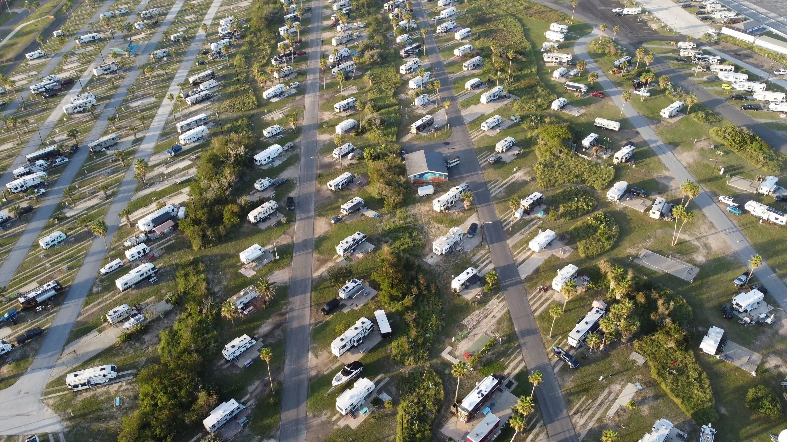 An aerial view of a RV park with numerous RVs parked on grassy sites, divided by roads and trees. There are some palm trees, small structures, and a paved parking lot visible.