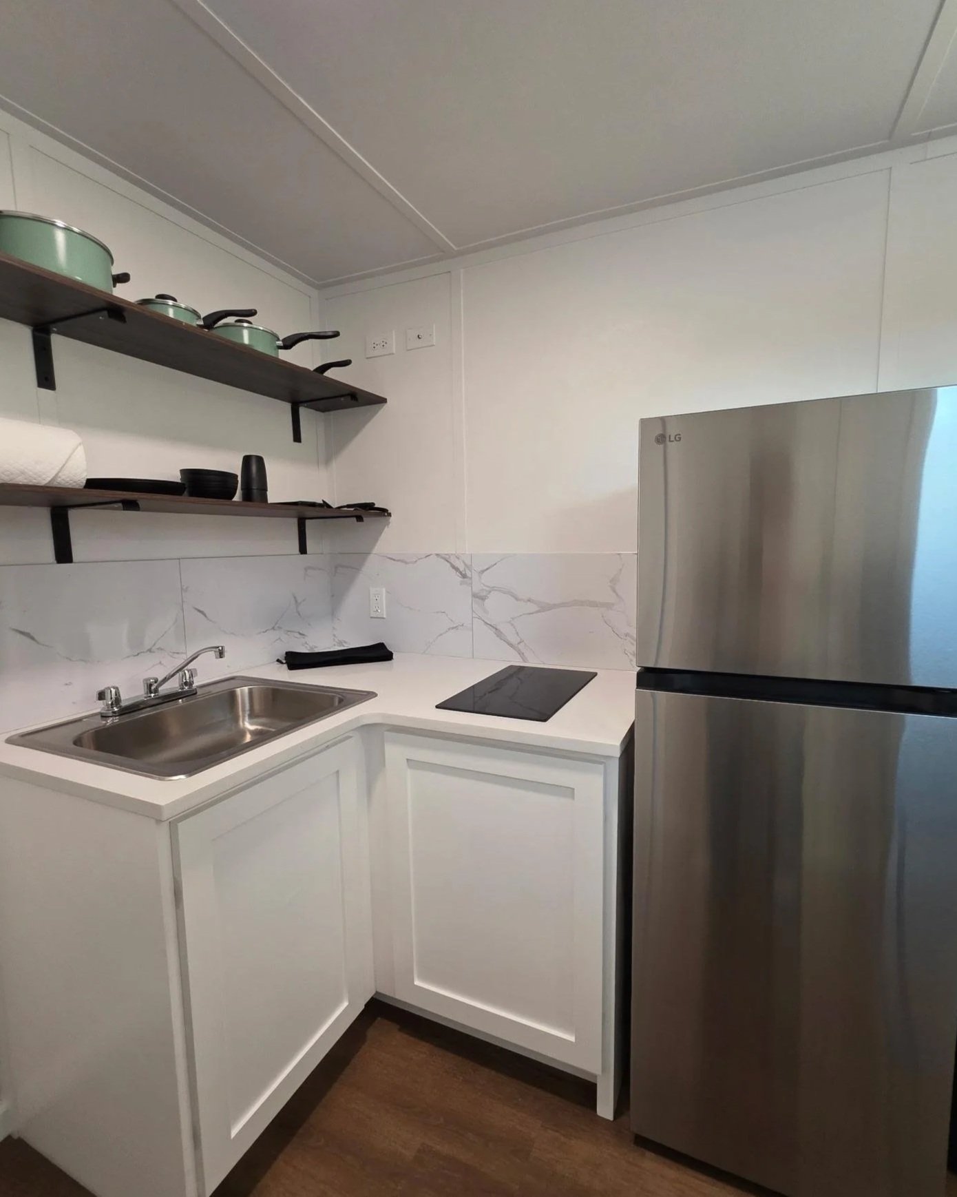 A modern kitchen with white cabinets, a metallic refrigerator, a black cooktop, a stainless steel sink, and floating shelves with pots and bowls.