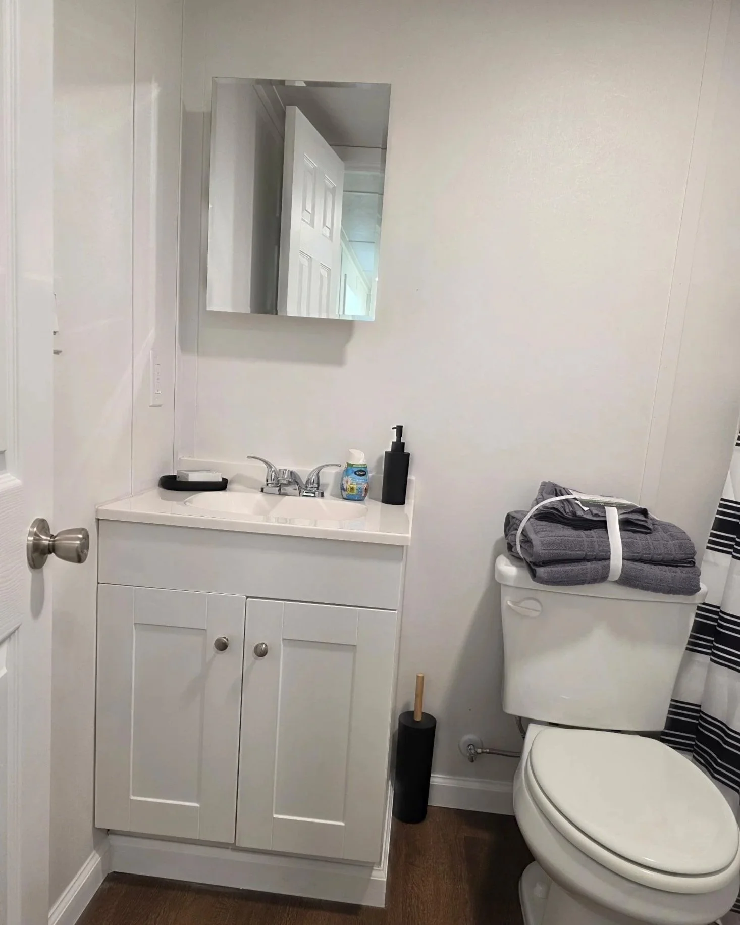 Bathroom with white vanity, mirror, small sink, black soap dispenser, toilet with gray and black striped towel on top, black toilet brush, and some folded clothes on the toilet tank.