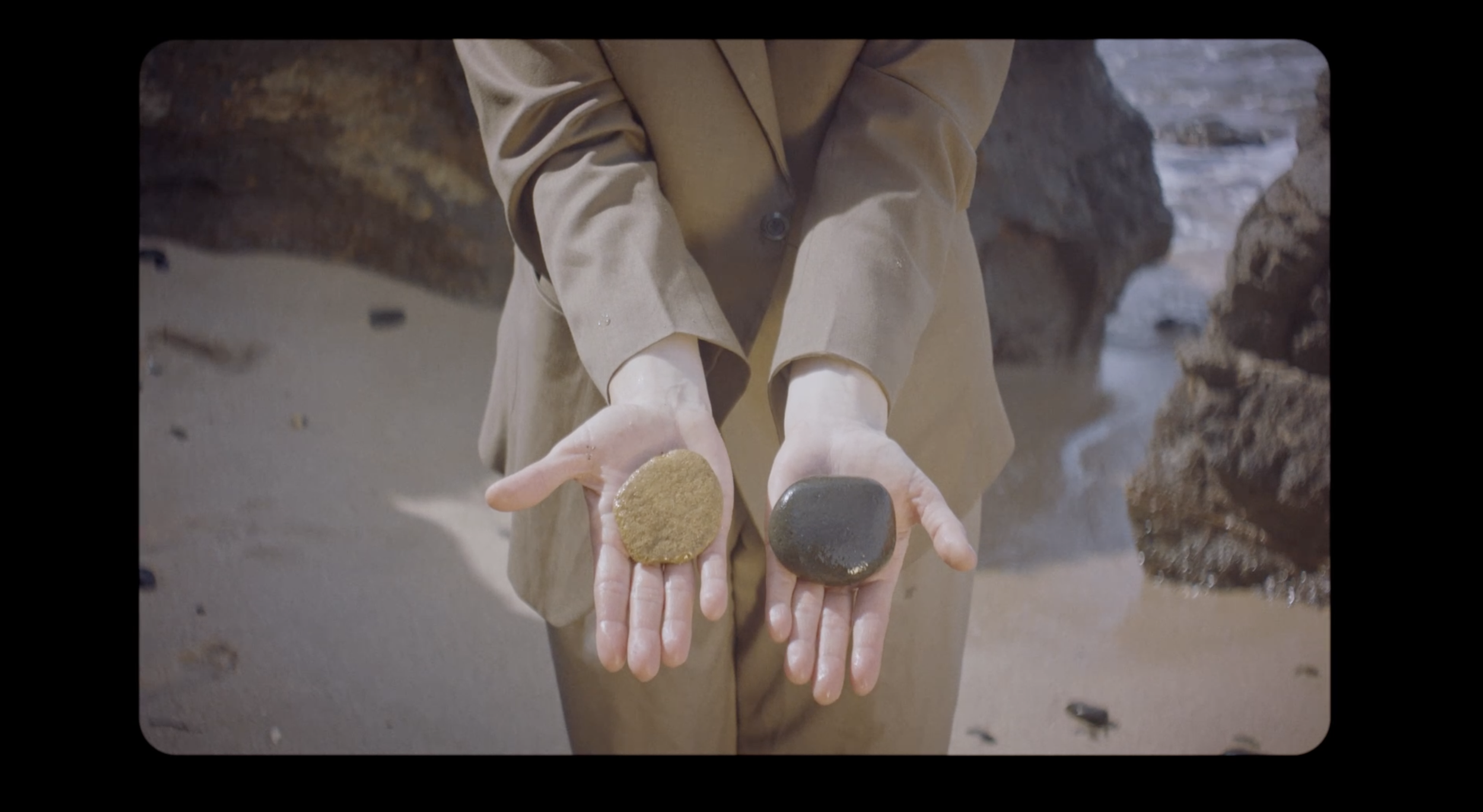 Person holding two circular objects, one beige and one dark, on a sandy and rocky beach.