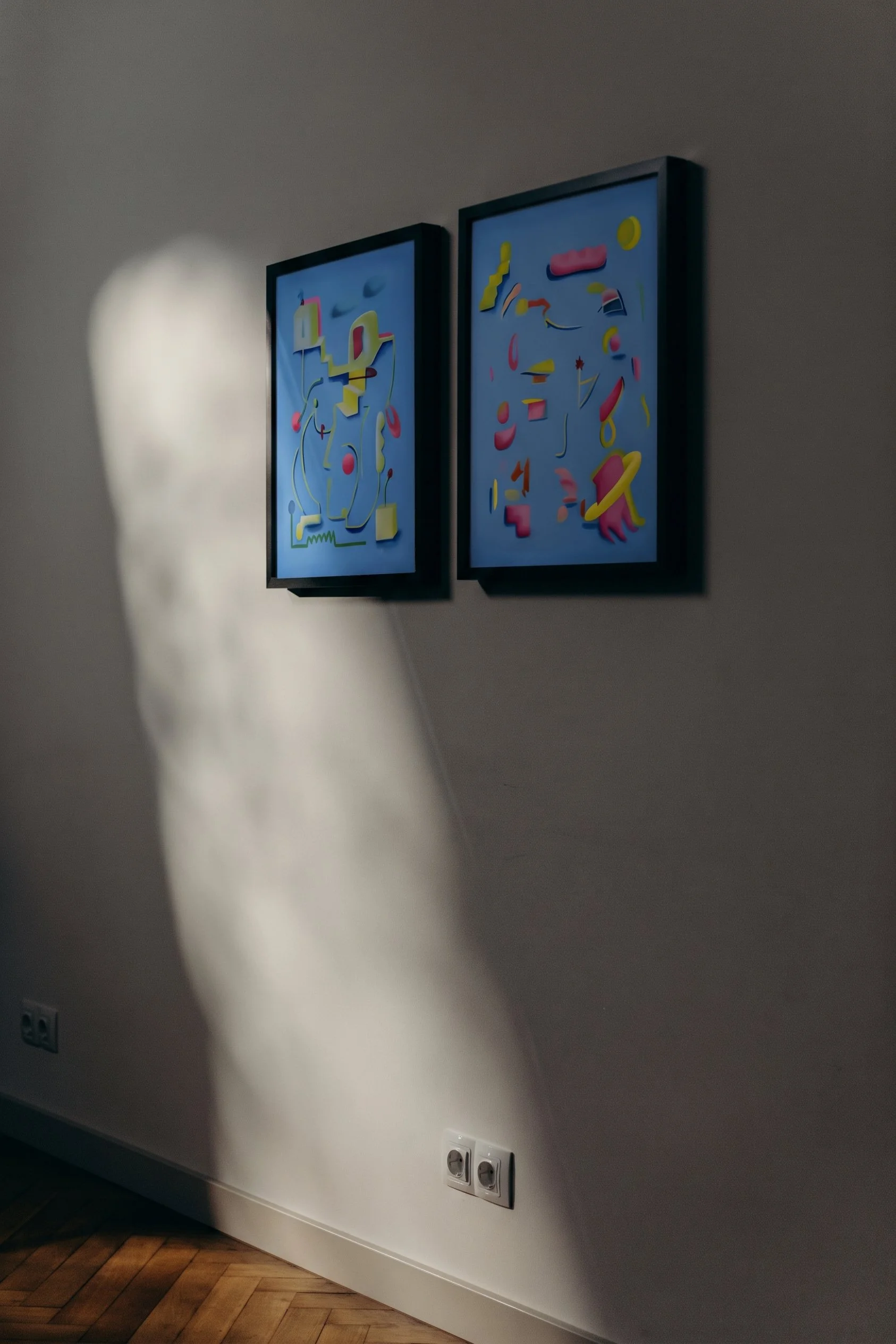 Two framed abstract digital art pieces hanging on a white wall, illuminated by a spotlight, with a white electrical outlet and a wooden floor visible at the bottom.