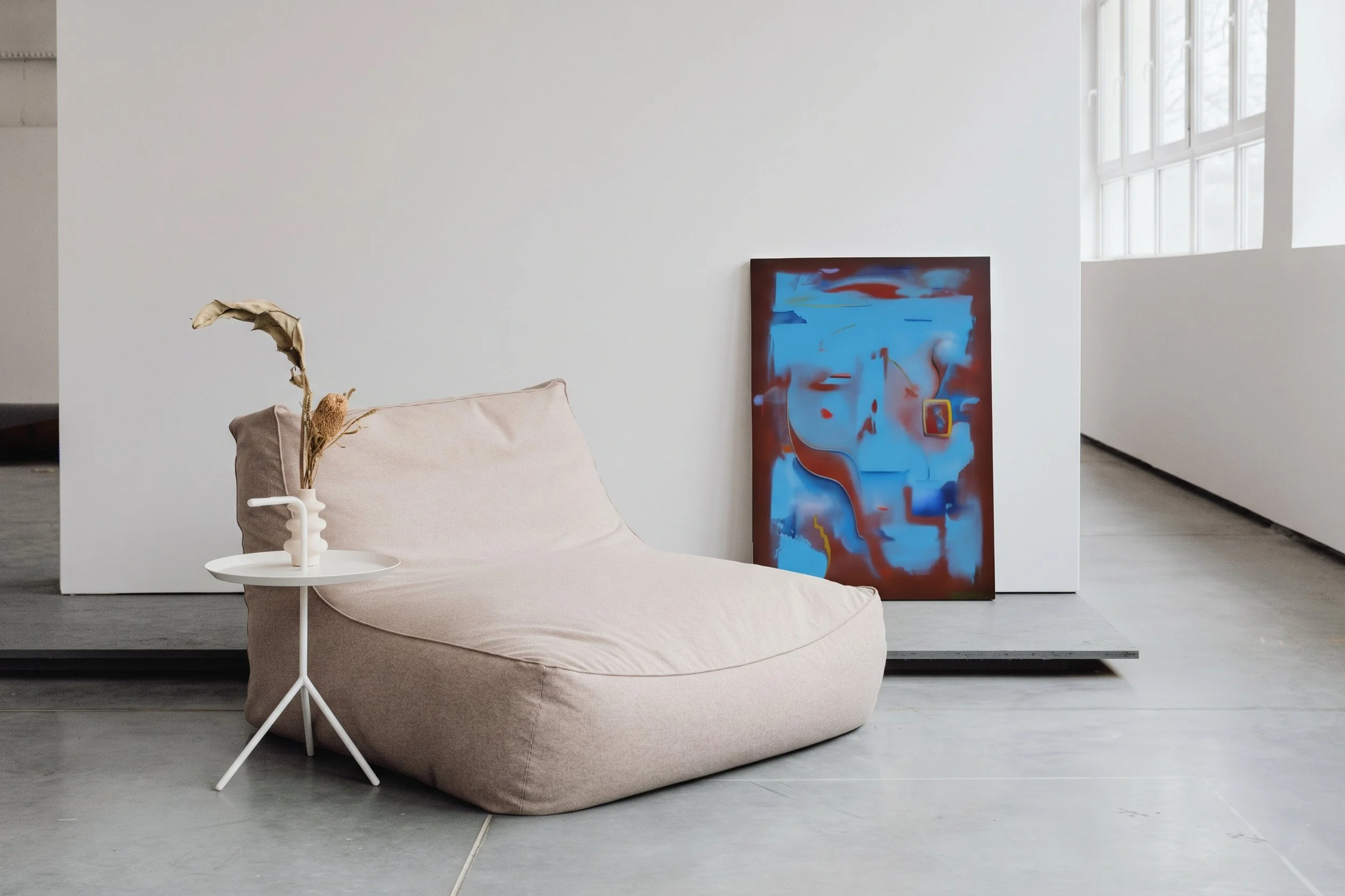 Minimalist interior with a beige cushioned lounge chair, a small white side table holding a dried plant arrangement, and an abstract painting with blue, red, and yellow colors resting against a white wall near windows.