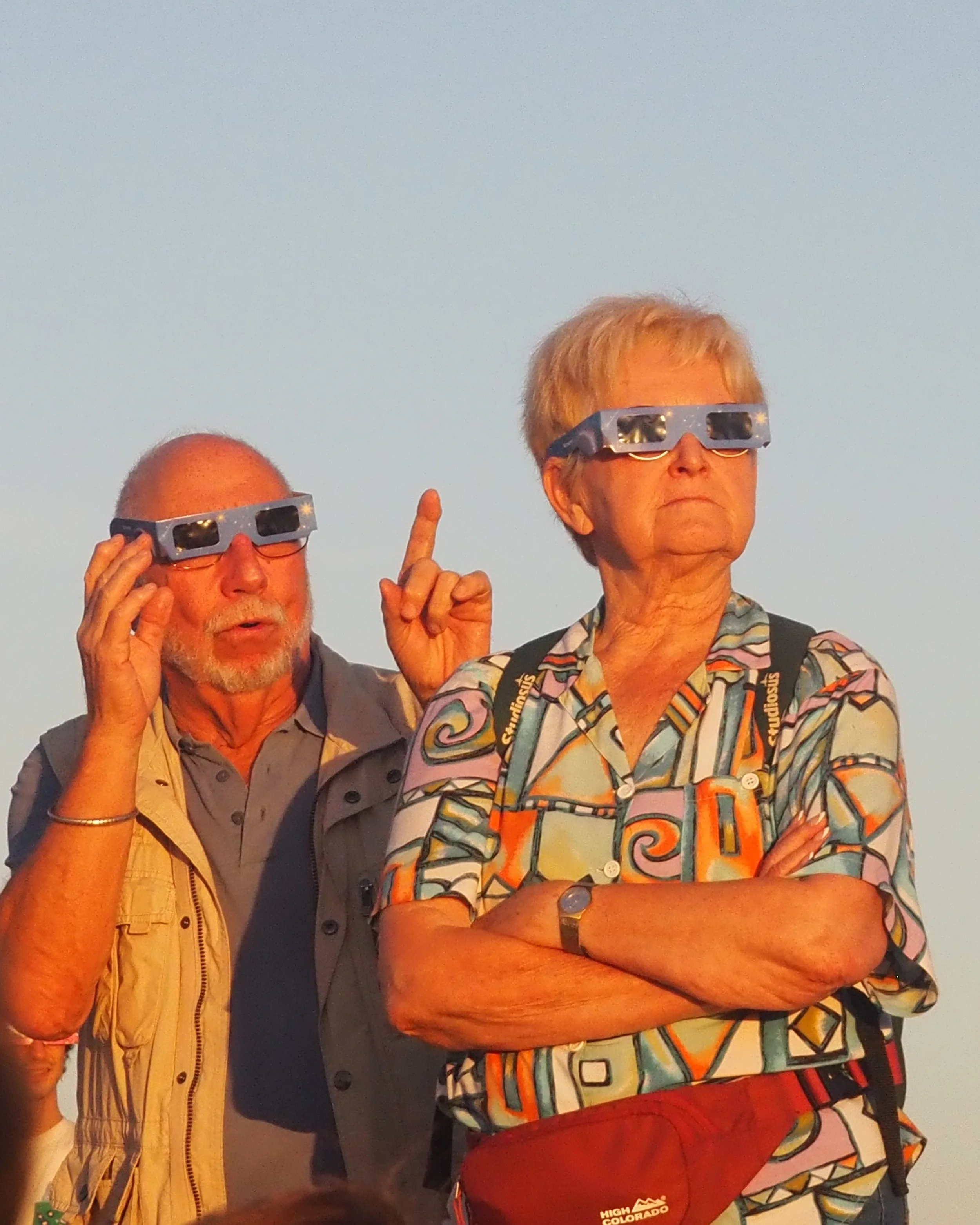 Two older adults, a man and a woman, wearing 3D glasses and standing outdoors against a clear sky. The woman has crossed arms, and the man is raising one finger with a serious expression.