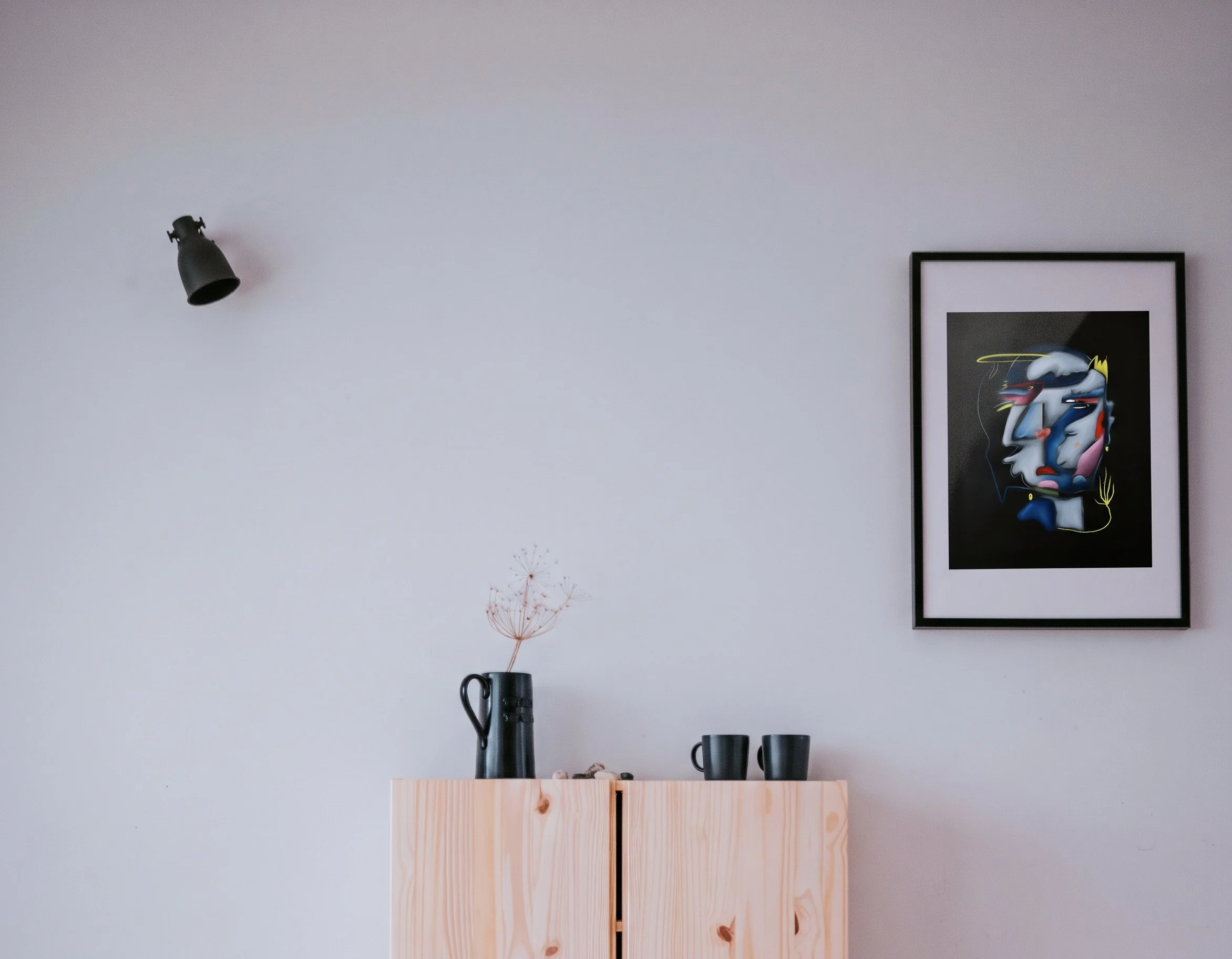 Interior of a room with minimal decor, including a black wall-mounted lamp, a framed colorful abstract artwork, and a wooden shelf with black vases and a vase with dandelion seed head, against a light-colored wall.