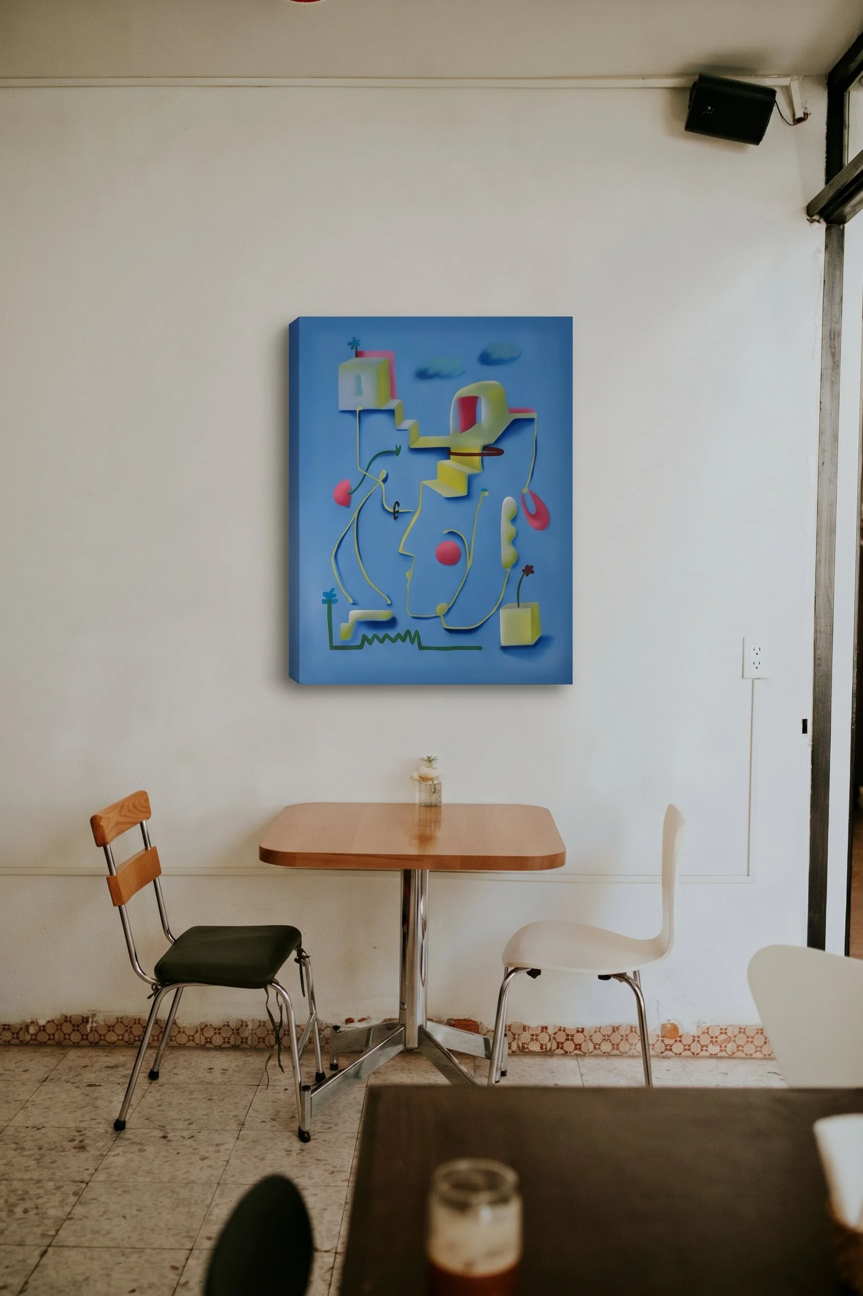 Interior of a room with a simple light-colored wall, featuring a colorful abstract painting. There are various chairs around a small wooden table, and a partial view of a glass of beverage on the table in the foreground.