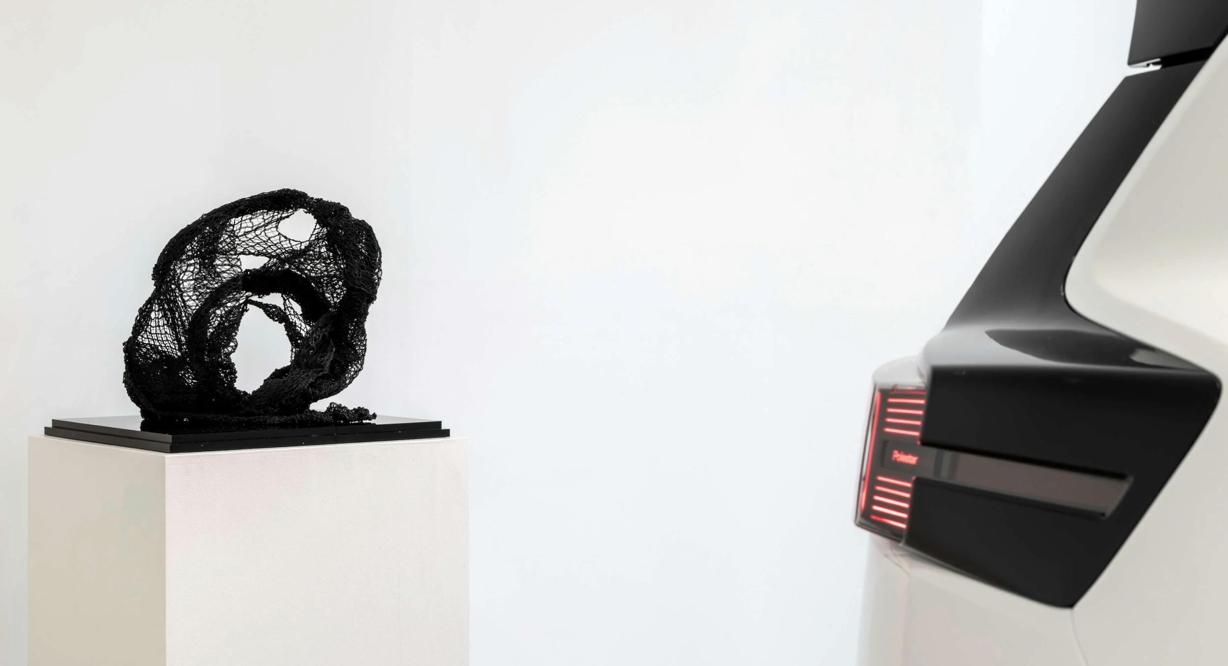 An abstract black sculpture with an intricate, mesh-like design displayed on a white pedestal, with part of a modern computer or device visible on the right side of the image.