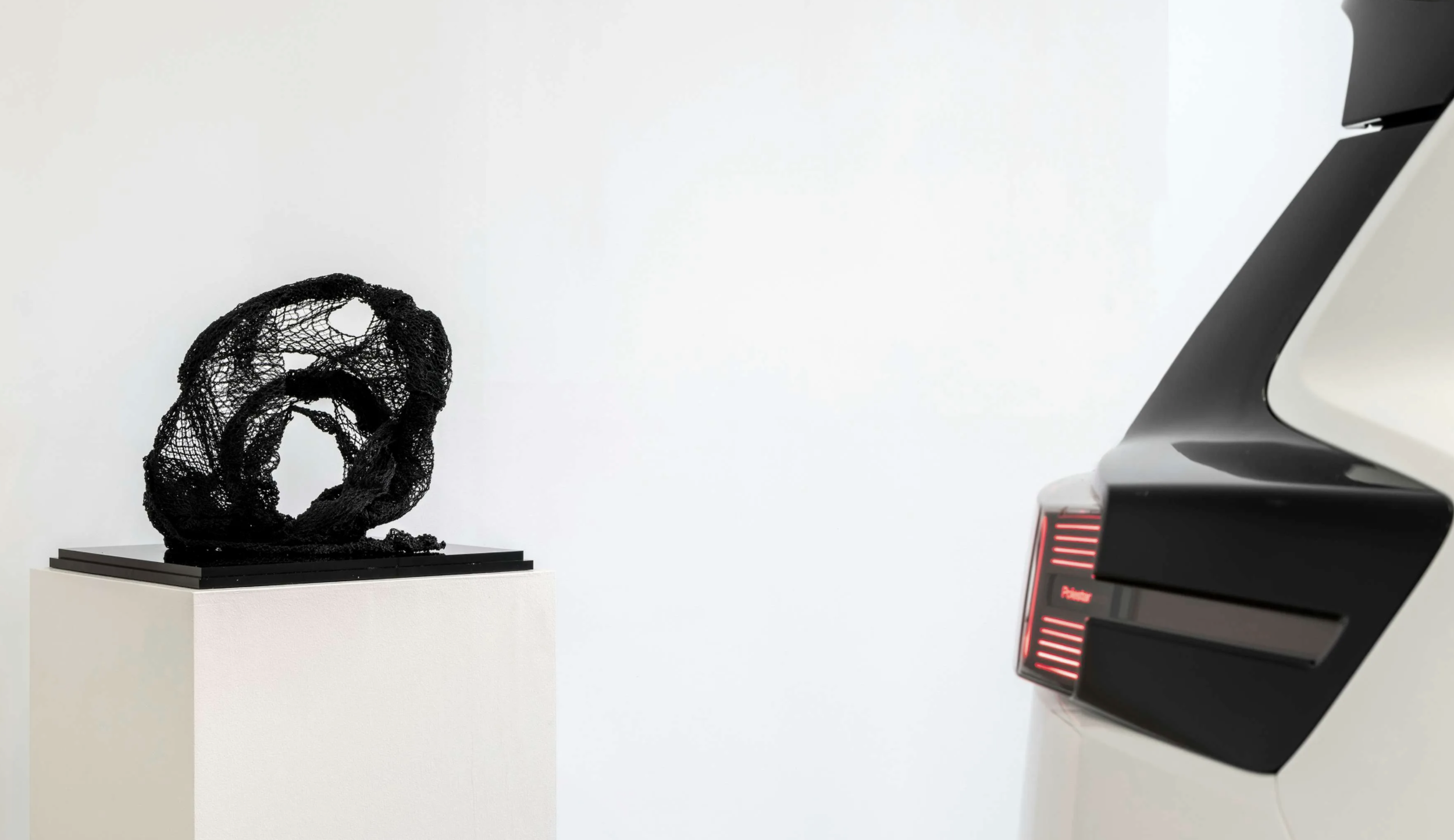 A black, sculptural art piece with an intricate, web-like structure on a white pedestal, and part of a futuristic electronic or machinery device with red lights on the right side.
