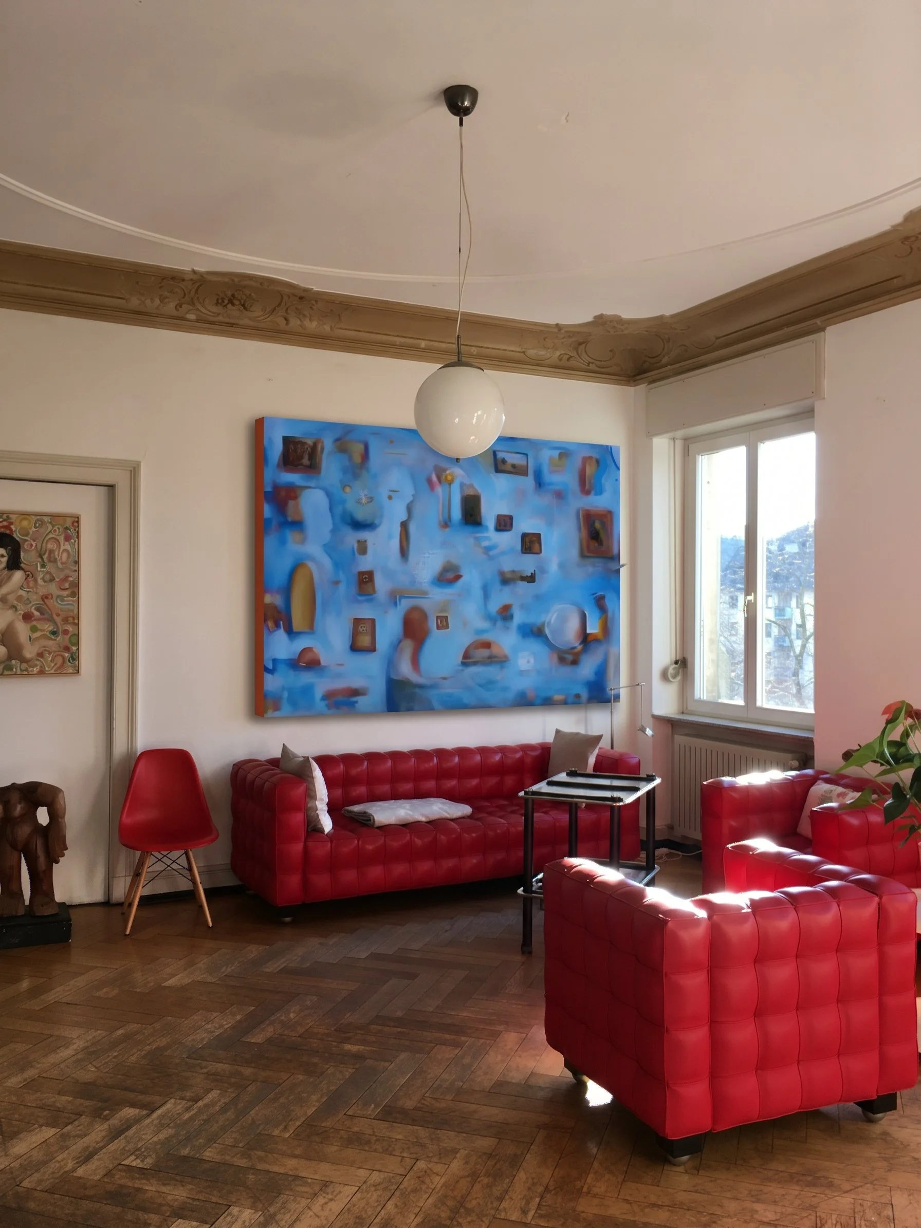A living room with a red leather sofa, a matching armchair, and a small black side table. The walls are decorated with colorful artwork, including a large abstract painting with blue and various shapes, and a smaller framed piece. There is a window with white frames allowing natural light to illuminate the room. The ceiling has ornate molding, and a spherical white pendant light hangs from the center of the ceiling.