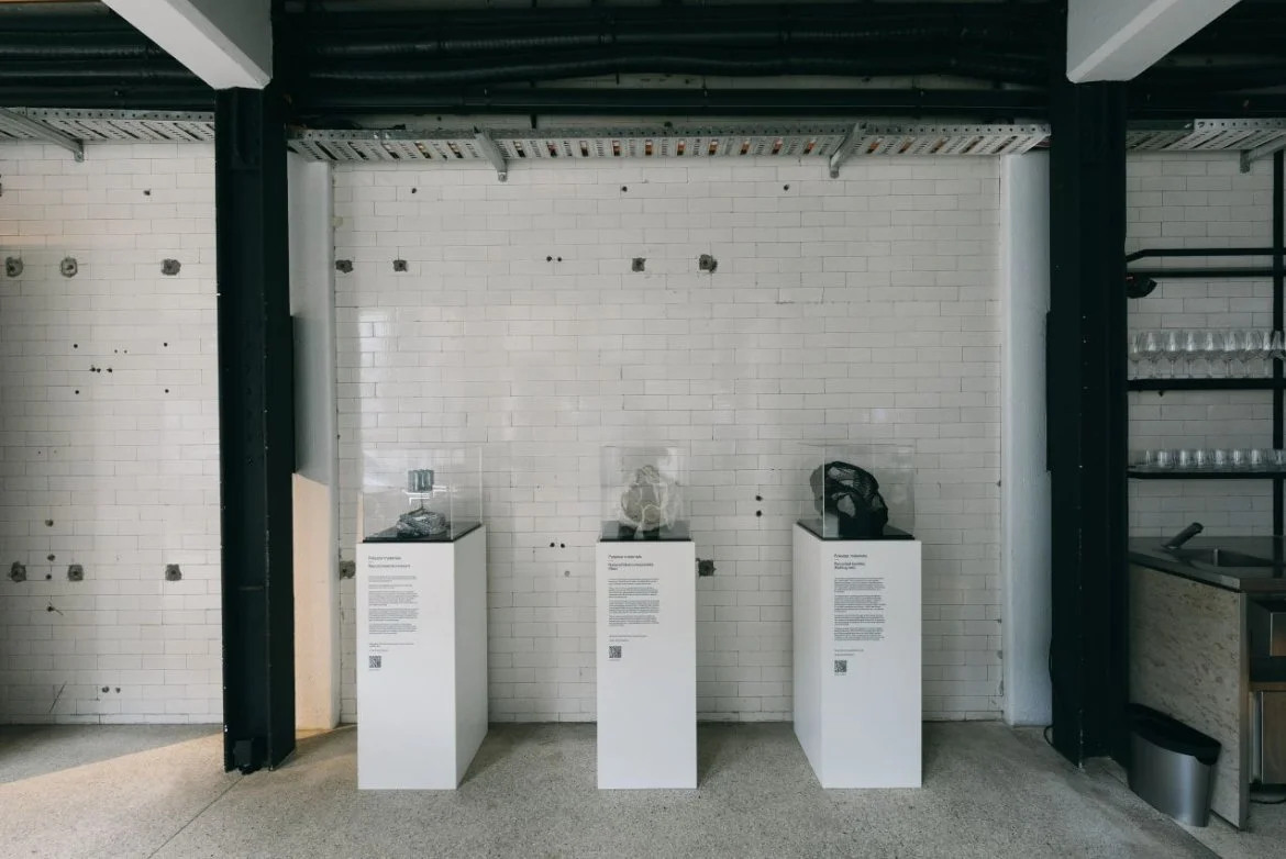 Three glass sculptures displayed on white pedestals against a white brick wall in a museum setting