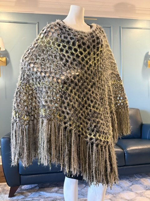 A multicolored, crocheted poncho with fringe hanging from the bottom, displayed on a mannequin in a room with dark blue walls and a black sofa.