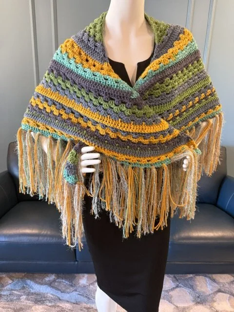 Mannequin wearing a colorful, crocheted shawl with fringe, featuring stripes of yellow, orange, green, gray, and blue, draped over shoulders and torso.