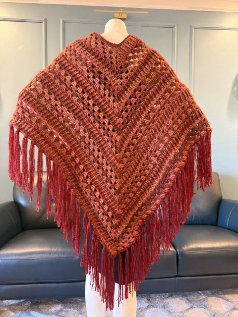 Red and brown crocheted shawl with fringe hanging on a mannequin in front of a blue wall and gray sofa.