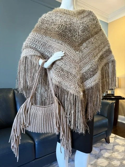 Mannequin dressed in a beige fringe poncho, holding a matching fringe handbag, in a living room with a sofa and lamp.