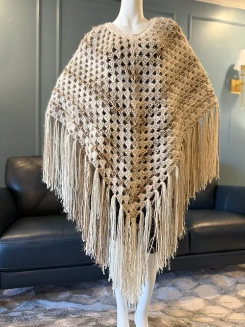 Mannequin wearing a large beige crochet shawl with fringe, displayed in a room with blue walls and black leather furniture.
