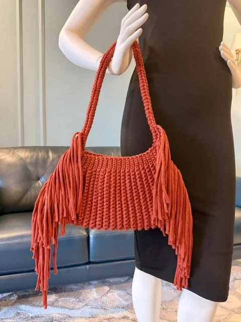 Red macrame handbag with fringes, held by a mannequin dressed in a black outfit, in a room with a leather couch and a doorway.