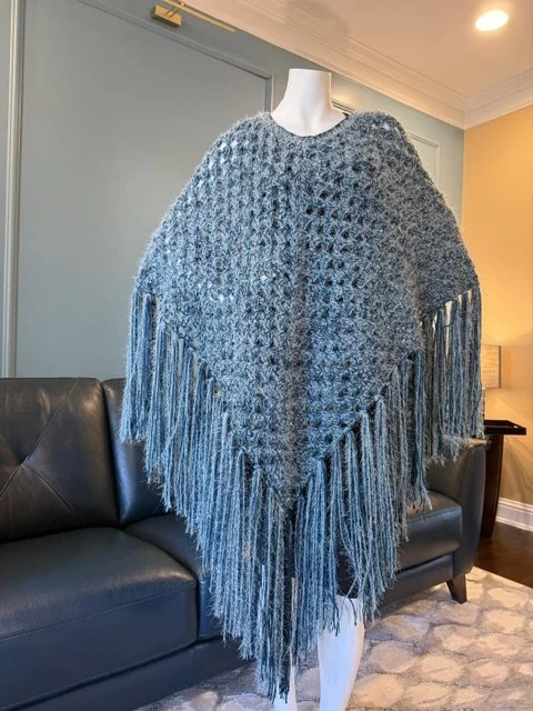 A mannequin wearing a blue knitted poncho with fringe details, displayed in a living room with a gray sofa and a lit lamp in the background.