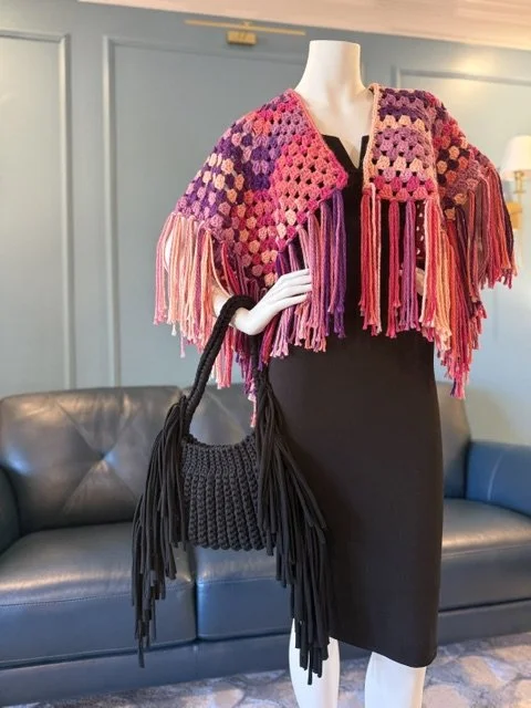 Mannequin wearing a colorful crochet shawl with fringe, a black dress, and holding a black fringe handbag in a room with blue walls and leather couches.