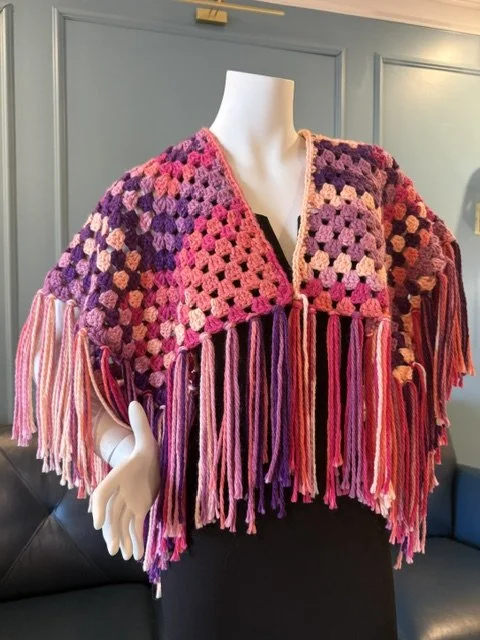 Colorful crochet shawl with fringe in pink, purple, and peach tones displayed on a mannequin.