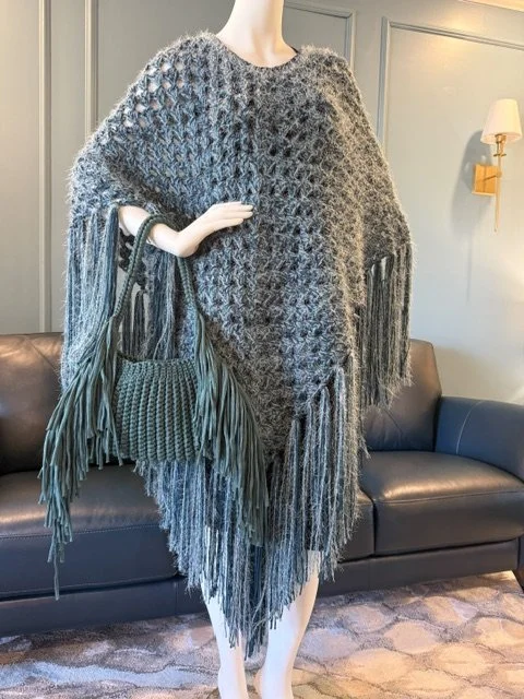 Person wearing a thick, textured, two-tone grey crochet poncho with long fringe details on the edges, carrying a large textured bag with fringe accents, standing in a stylish living room.