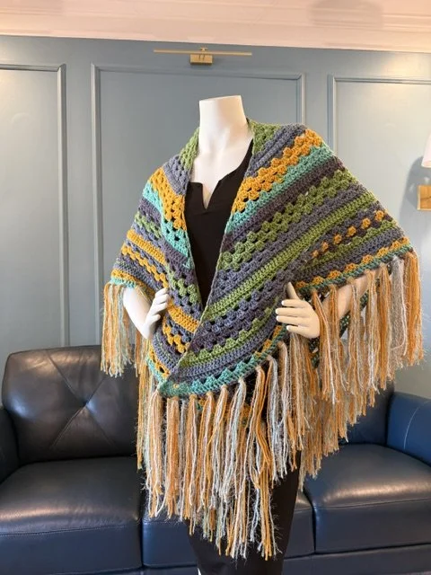 A mannequin wearing a multicolored crocheted shawl with fringe, draped over shoulders, and a black outfit underneath, set against a blue paneled wall with a black leather sofa nearby.