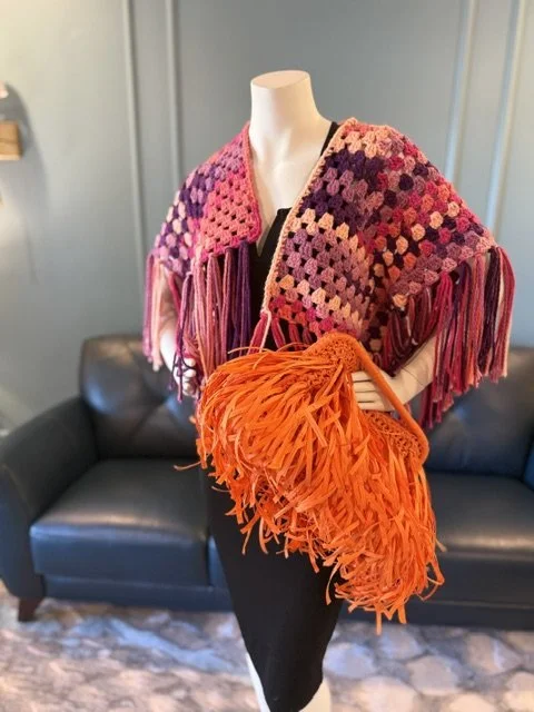 Colorful crocheted shawl with fringe draped over a mannequin in a room with a black leather couch.