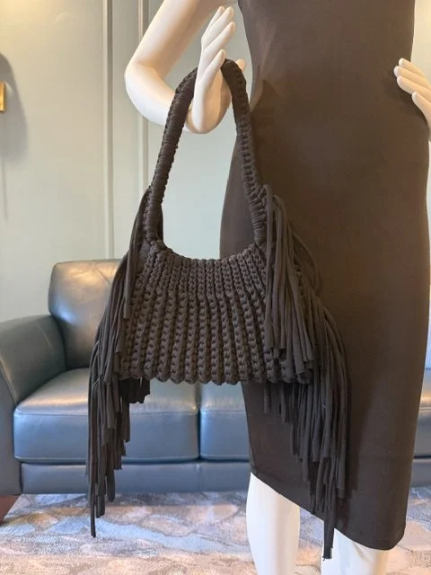 Mannequin holding a black, knitted handbag with fringe details, standing in a room with a grey couch in the background.