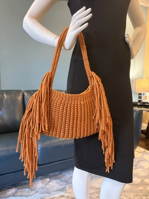 A mannequin wearing a black dress, holding an orange fringed knit handbag in a living room setting.