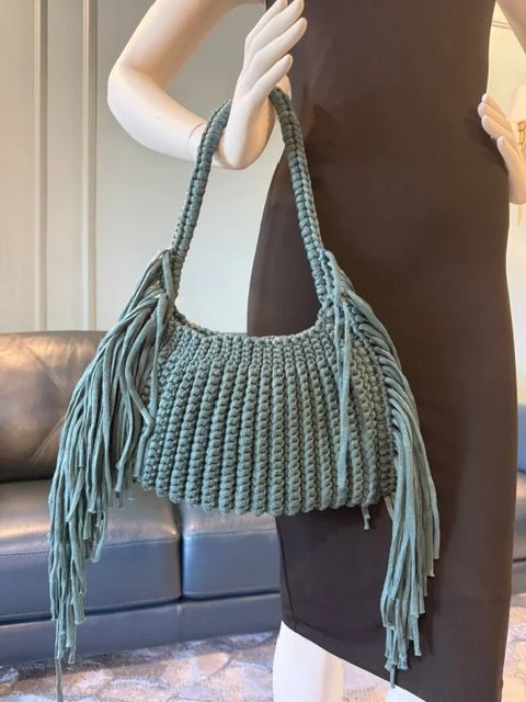 A green knit handbag with fringes hanging from the sides, held by a mannequin wearing a black dress.
