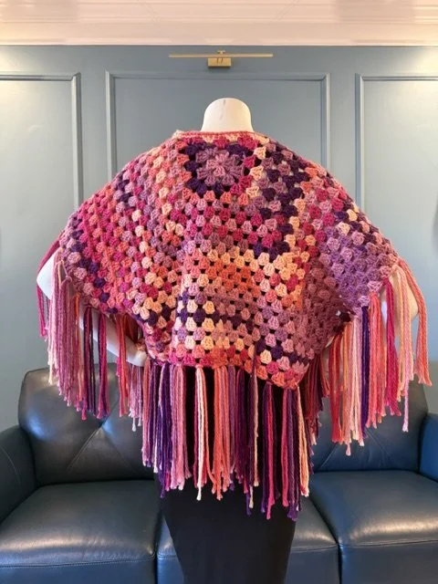 A mannequin wearing a colorful, granny square crochet shawl with long pink, purple, and red fringes hanging from the edges, displayed in front of a blue wall.