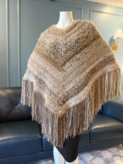 Beige knitted poncho with fringe details displayed on a mannequin in a living room.