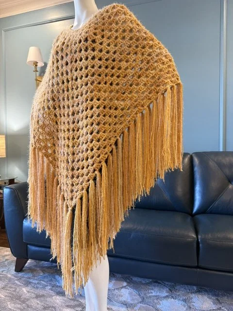 A mannequin wearing a tan crochet shawl with long fringe, displayed in a room with a black leather sofa, wall lamp, and blue wall.