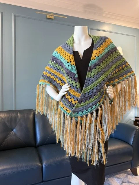 Mannequin wearing a colorful, multicolored, crocheted shawl with fringe, over a black dress, inside a room with blue walls and a black leather sofa.