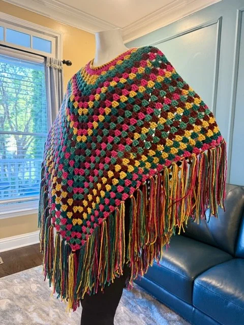 Colorful crocheted shawl with a fringe of multicolored yarns displayed on a dress form in a bright room.