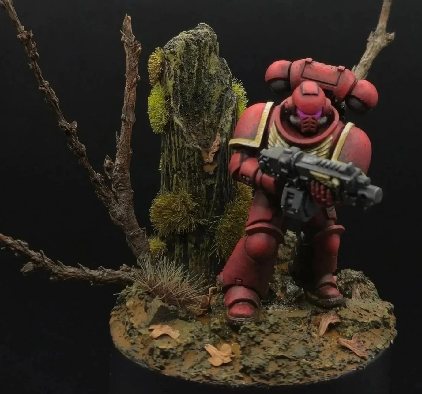 Figurine de soldat en armure rouge tenant une arme, posée devant un arbre fané avec de la mousse et des branches, fond noir.