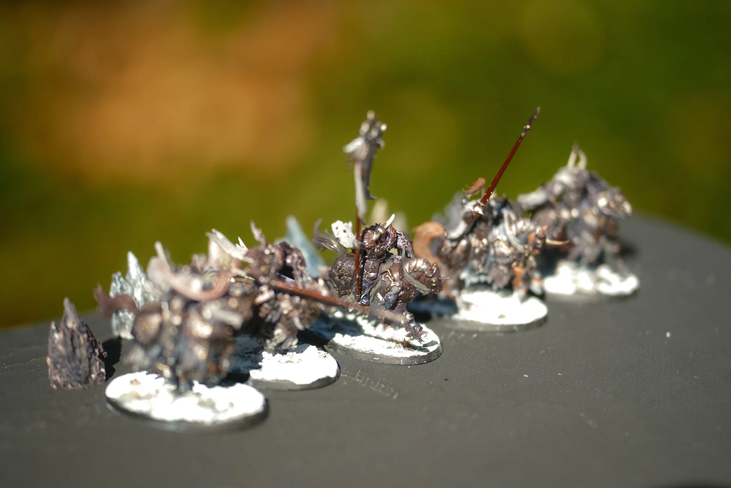 Miniatures de soldats en métal peint, alignés sur une surface noire, avec un fond flou de couleurs vertes et brunes.