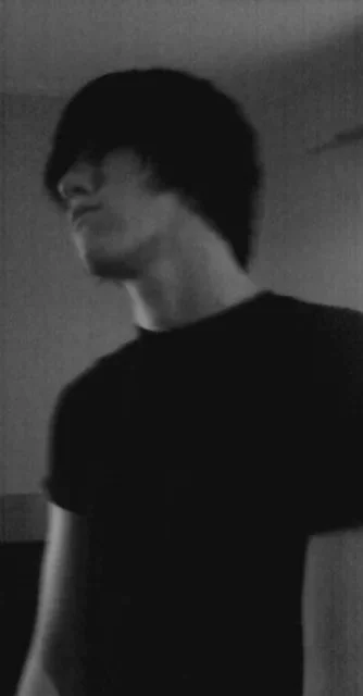 Un jeune homme avec les cheveux noirs et longs, portant un t-shirt noir, pris en selfie dans un miroir