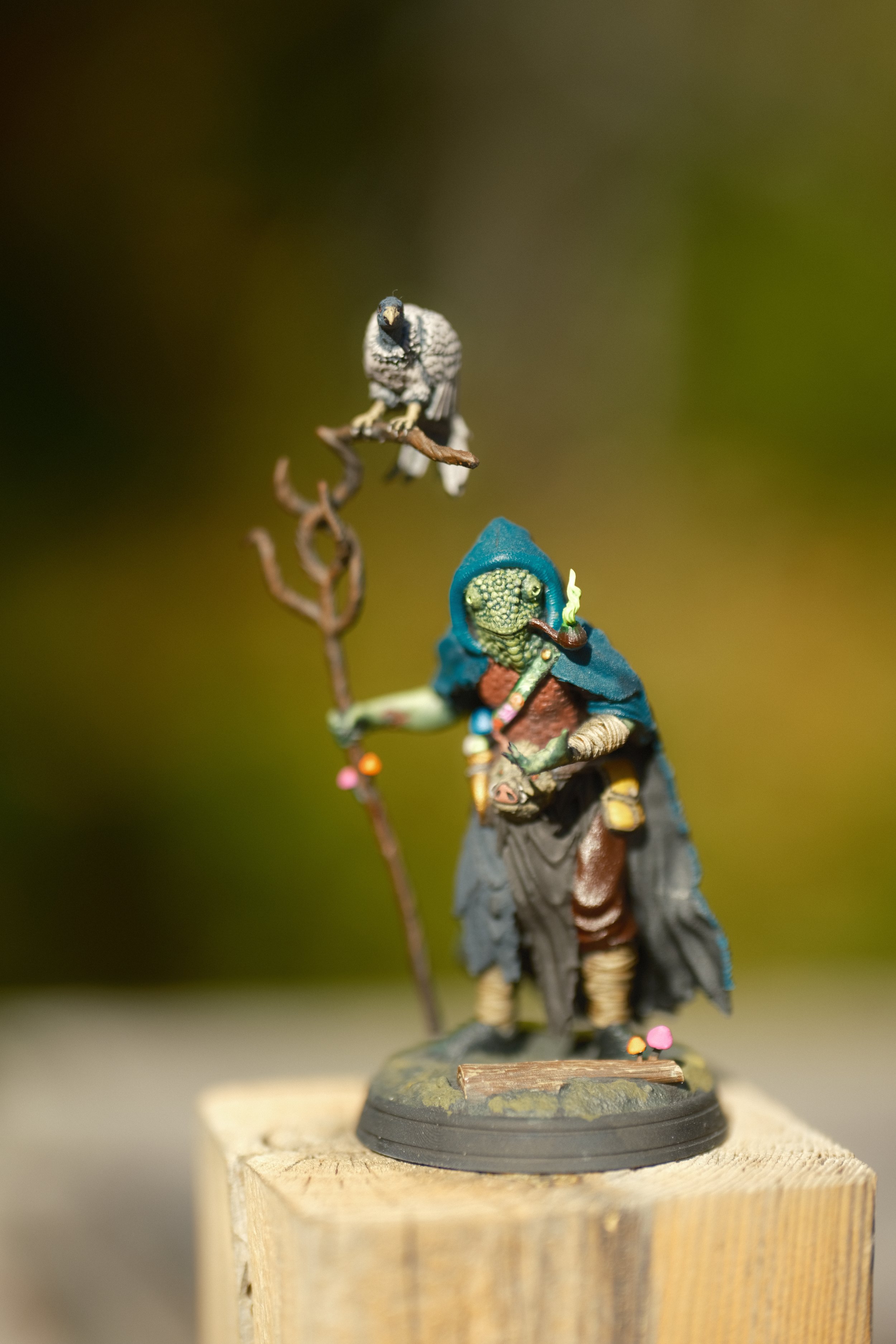 Petite figurine d'un personnage fantastique avec un chapeau bleu, une cape, et tenant un bâton, placée sur un socle en bois.