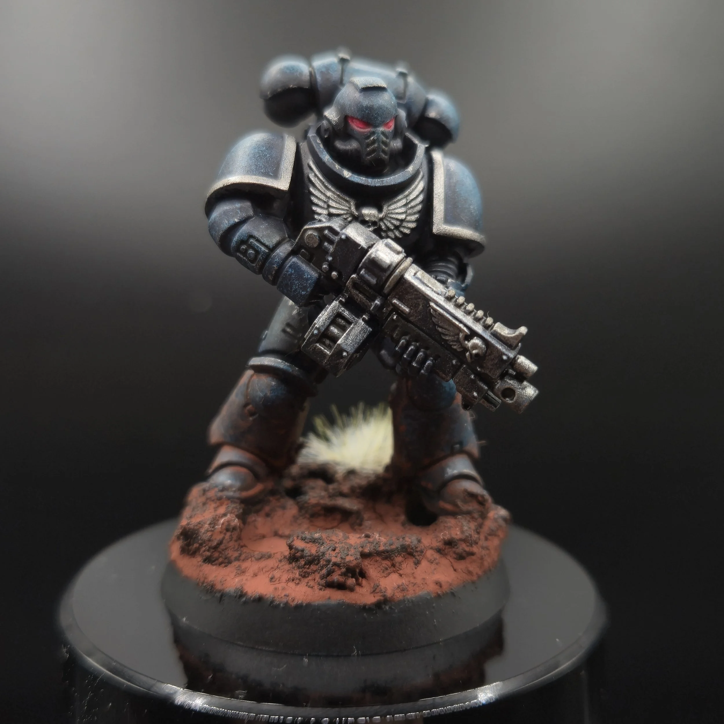 Une figurine d'un soldat en armure sombre avec des yeux rouges, tenant un arme futuriste, sur une surface rocheuse rougeâtre, contre un fond gris.