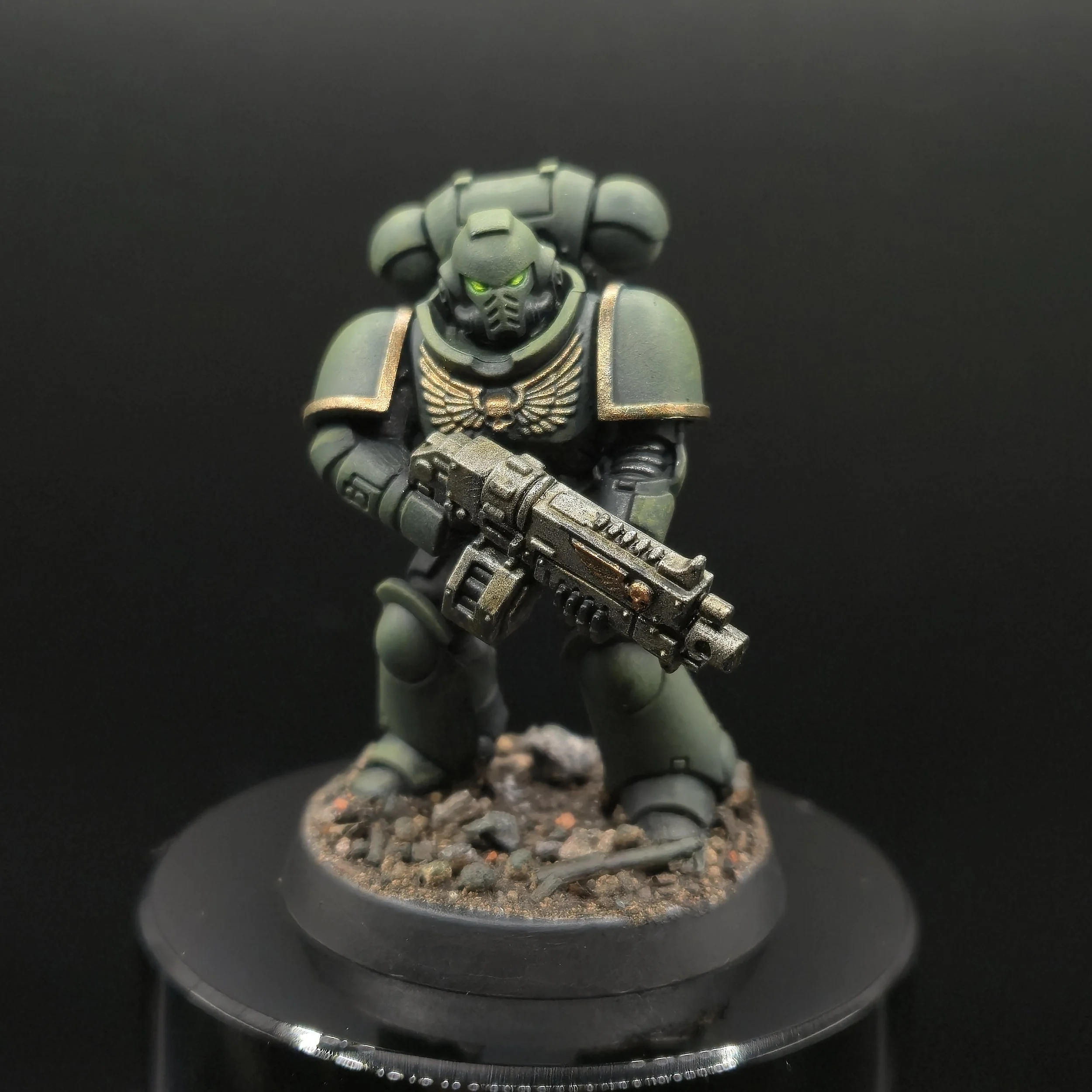 Figurine de soldat futuriste avec casque à visière verte, armure grisâtre, tenant une arme avancée, positionnée au-dessus de dures roches, sur un fond noir.
