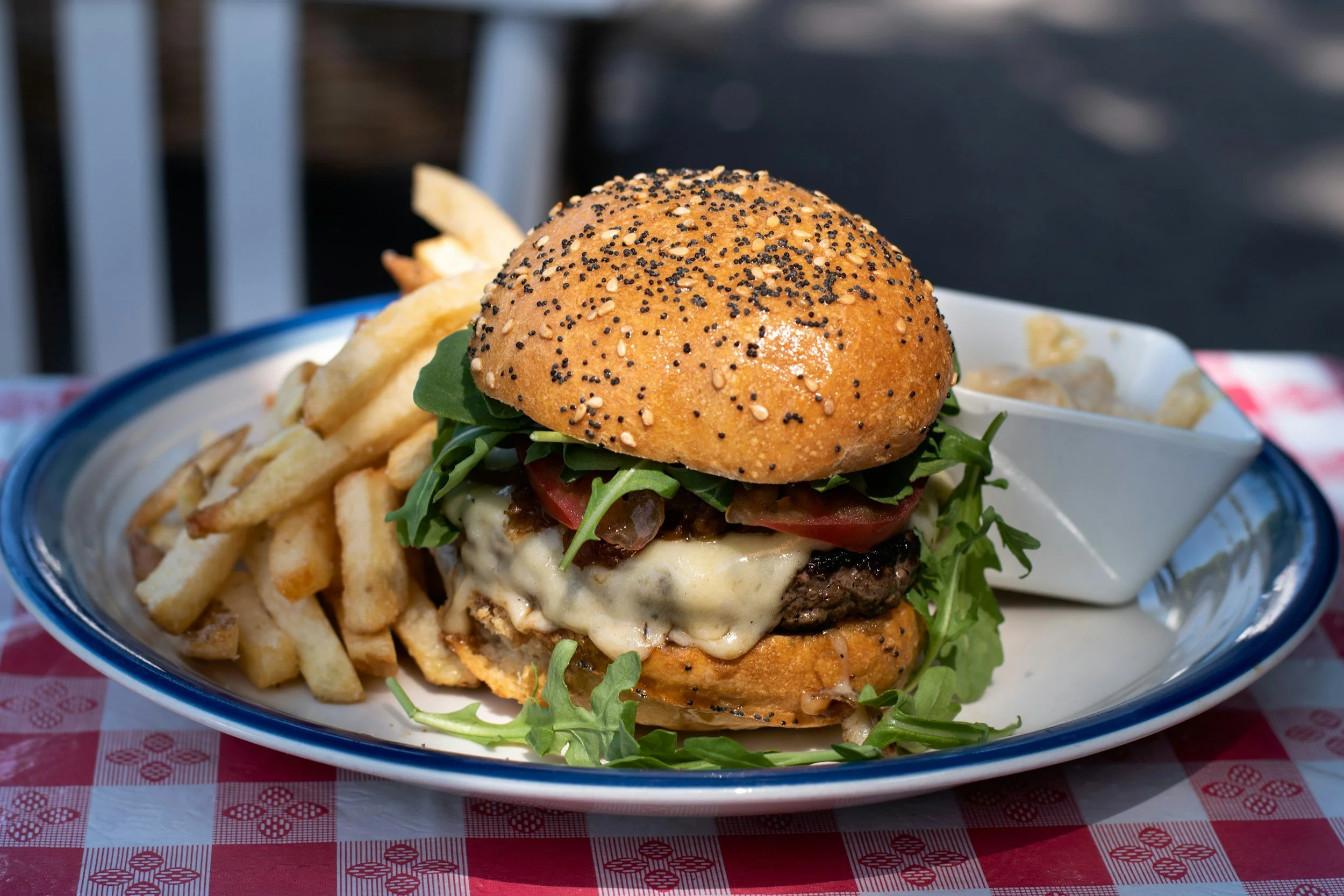 A cheeseburger with tomato, lettuce, pickles, melted cheese, and a sesame seed bun served with French fries on a white and blue plate on a red checkered tablecloth.