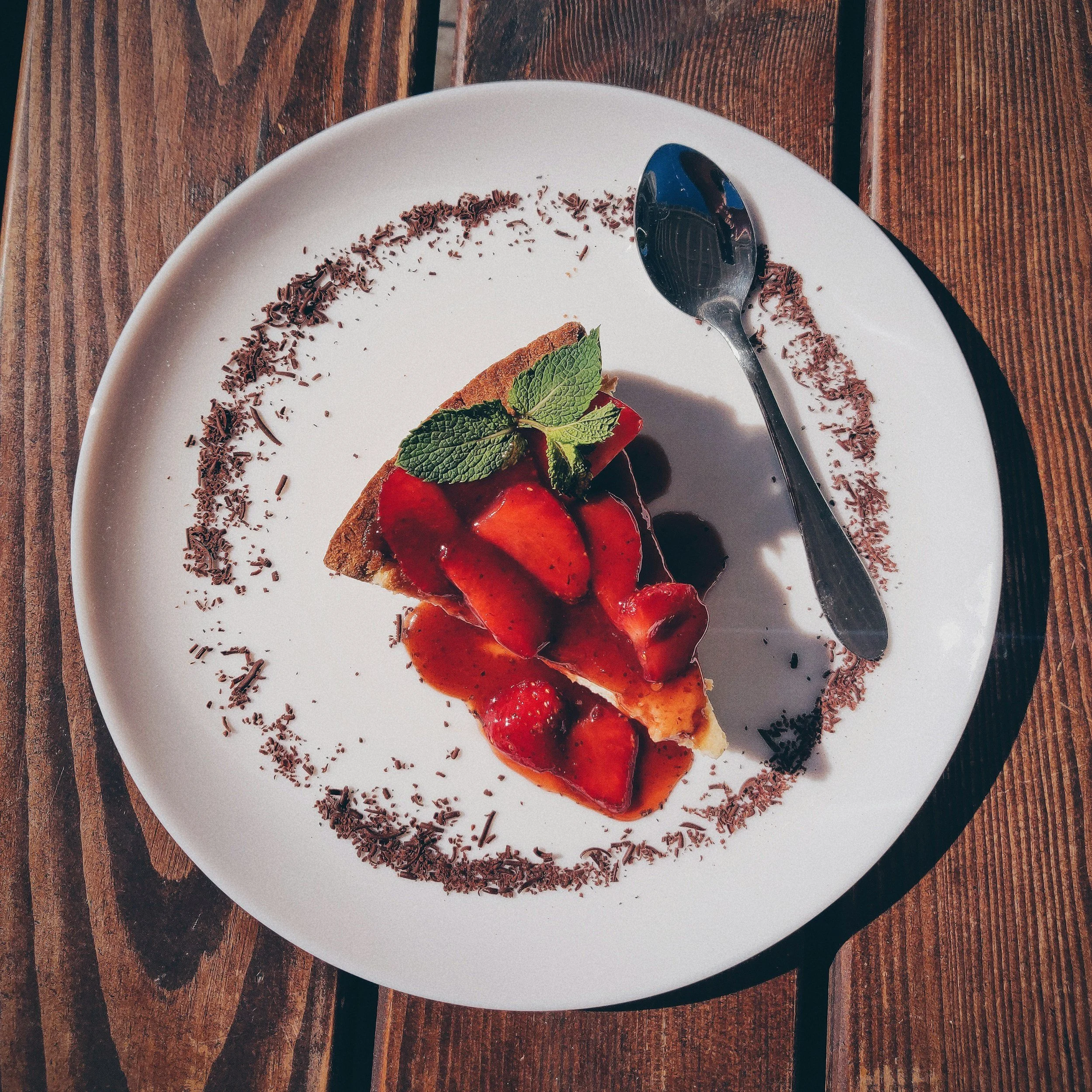 A white plate with a slice of chocolate cake topped with strawberries and a mint leaf. Chocolate shavings are scattered along the edge of the plate. A spoon rests on the plate, all on a wooden table.