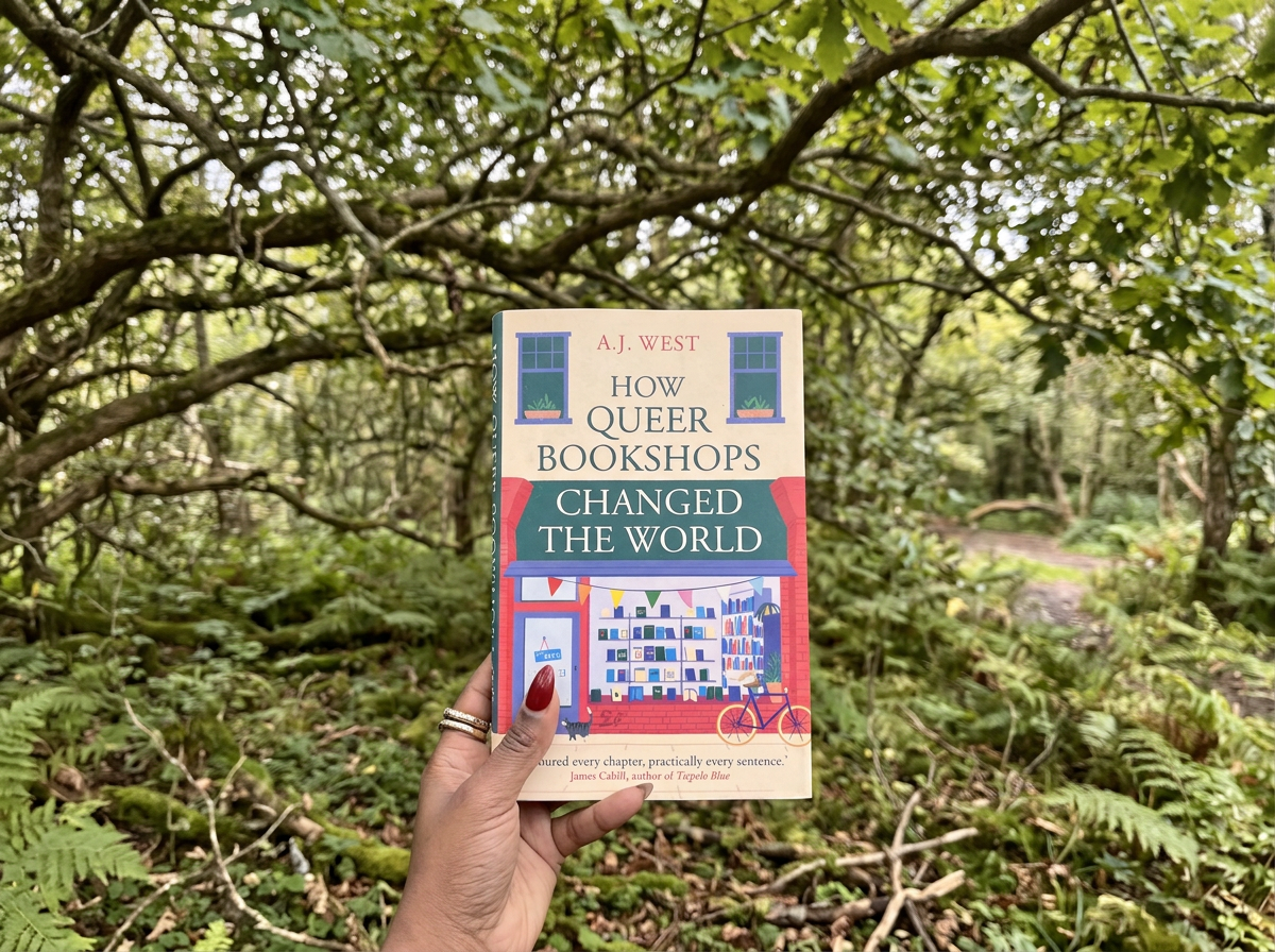 Hand holding a book titled 'How Queer Bookshops Changed The World' by A.J. West in a lush green forest with trees and branches overhead.