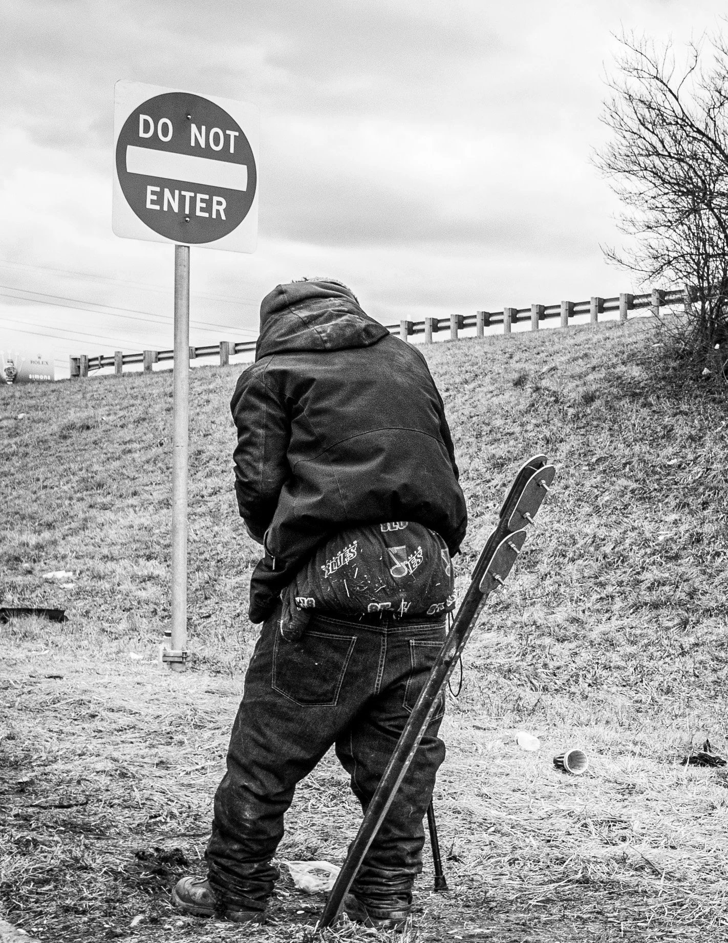 Black-and-white photograph of a person in a hooded jacket beneath a Do Not Enter sign beside a grassy embankment.