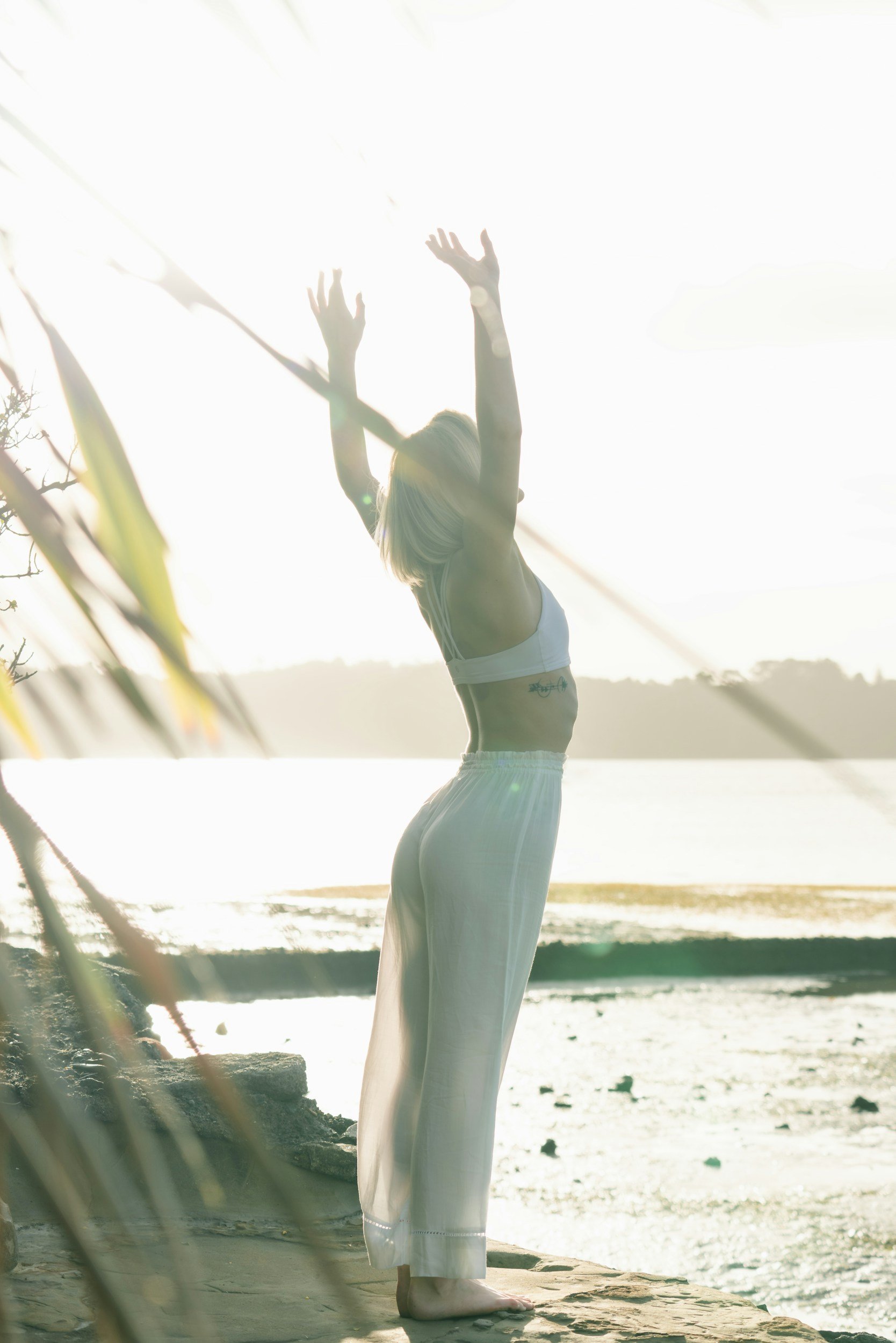 Frau beim Yoga am Strand während des Sonnenuntergangs, mit erhobenen Armen, Sonnenlicht im Hintergrund.