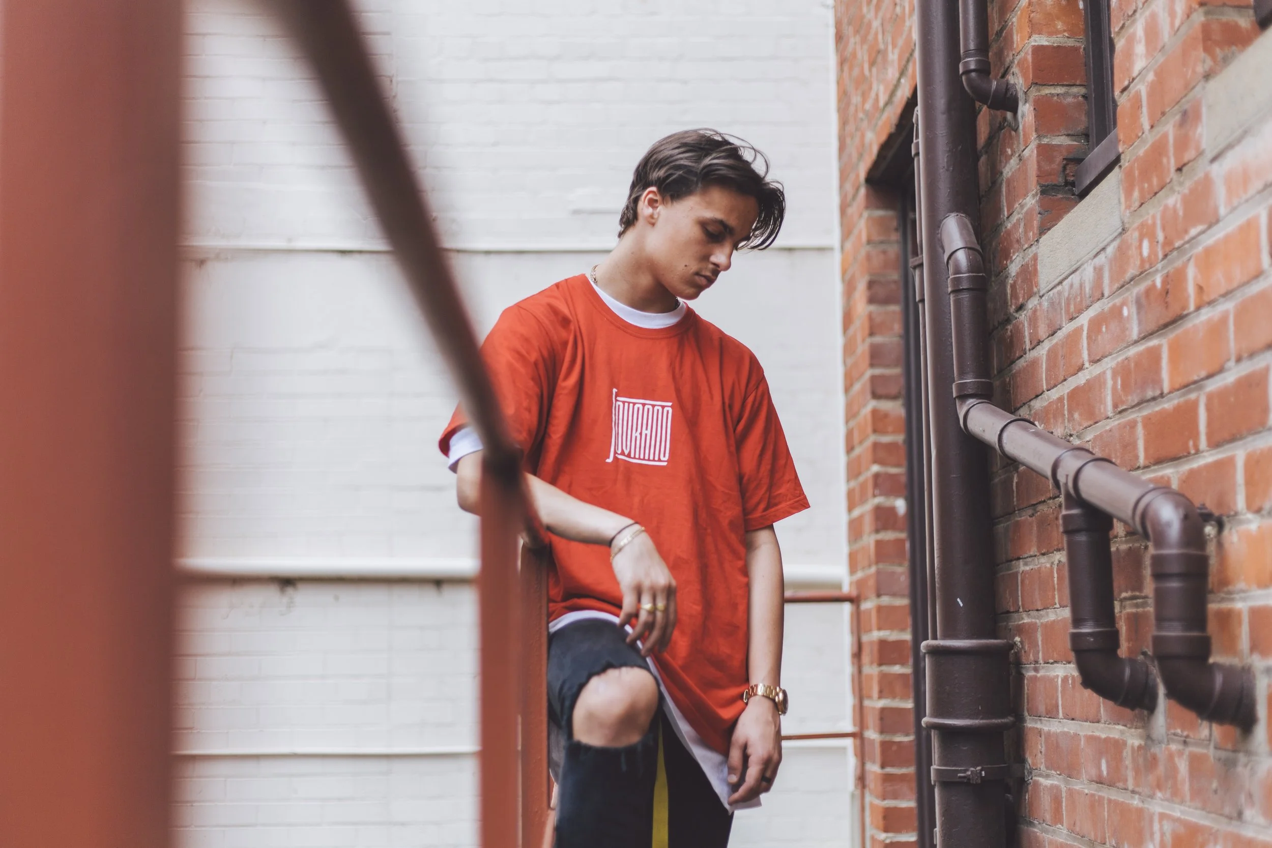 Two young men standing against a brick wall outside, one wearing a red t-shirt and black shorts, the other in a black distressed jean shorts with a white t-shirt underneath, both looking down with neutral expressions.