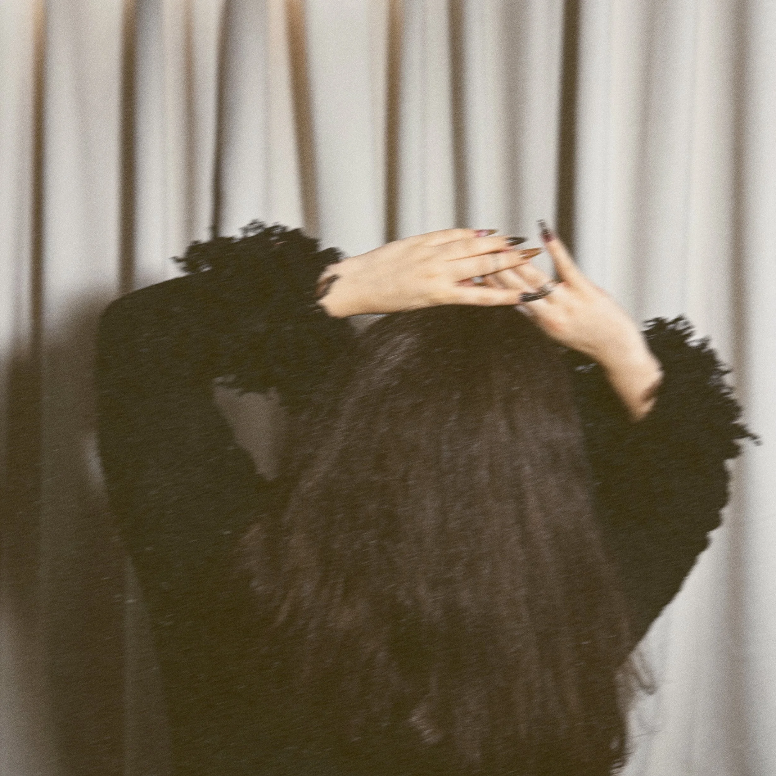 Close-up of person's hands with long dark nails, wearing black lace sleeves, clasped together, background of beige and brown striped curtain.